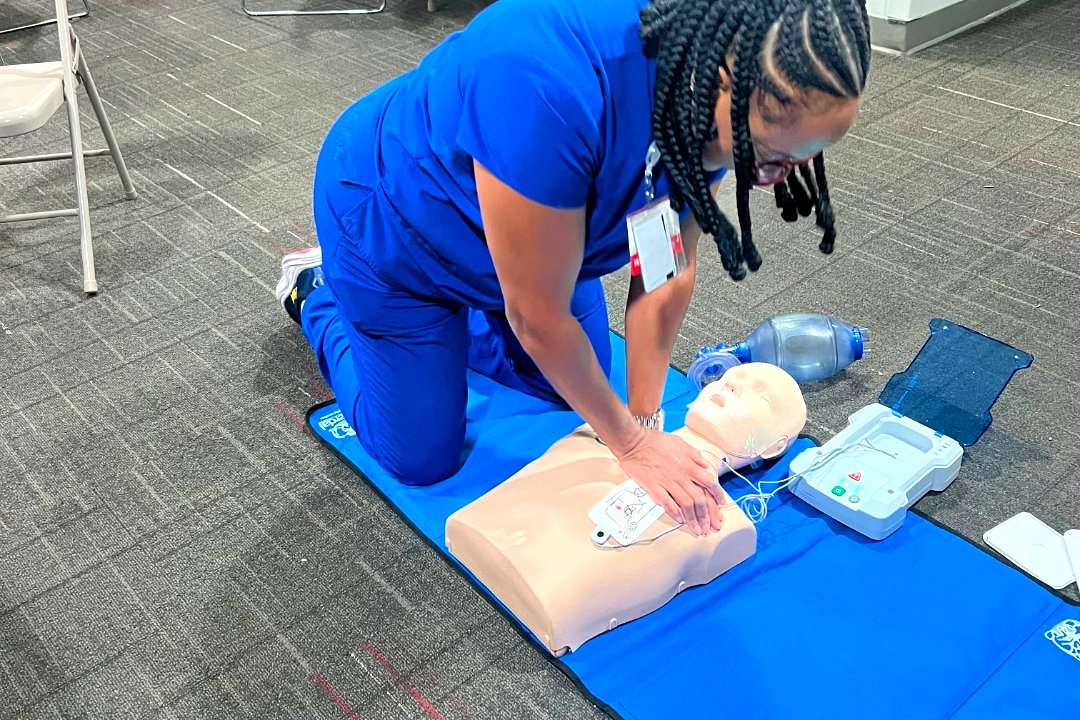 Nurse in blue scrubs performing chest compressions on an adult CPR manikin with an AED trainer beside her