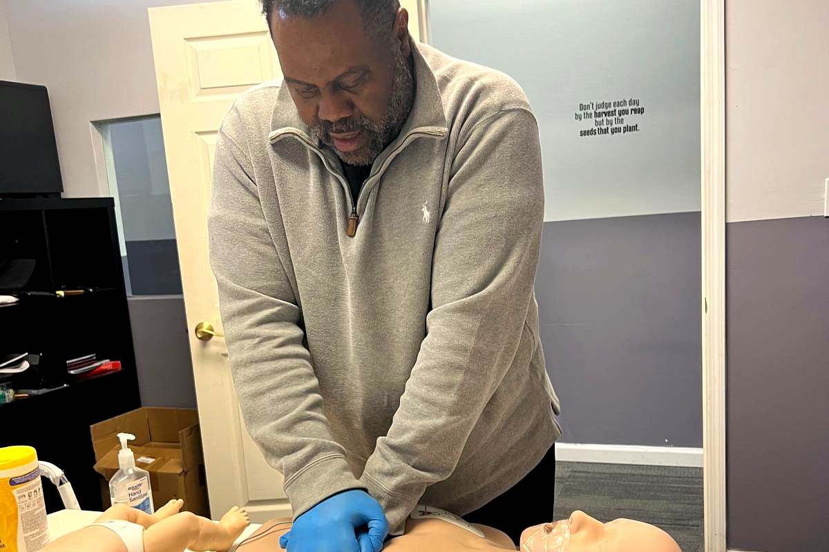 Student practicing chest compressions on an adult manikin at a classroom table with an AED trainer nearby